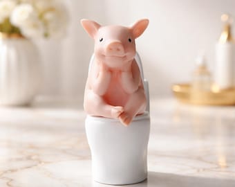 Op de afbeelding: Een roze varkensbeeldje dat op een witte toiletpot zit. Het varken heeft zijn handen onder zijn kin en een glimlachende uitdrukking. Het is een decoratief item.
