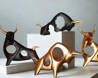 Op de afbeelding: Een verzameling decoratieve stierensculpturen in zwart en goud. De stieren hebben een cirkelvormige opening in hun lichaam en goudkleurige hoorns. De sculpturen worden tentoongesteld op een wit oppervlak tegen een lichtgrijze achtergrond.