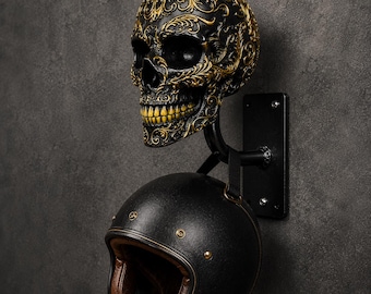 Op de afbeelding: Een wandhouder voor helmen met een zwarte schedel voorzien van gedetailleerde gouden filigraanversieringen. De schedel is bevestigd aan een zwarte metalen beugel die naar buiten steekt en een zwarte motorhelm met gouden klinknagels en bruine voering ondersteunt.