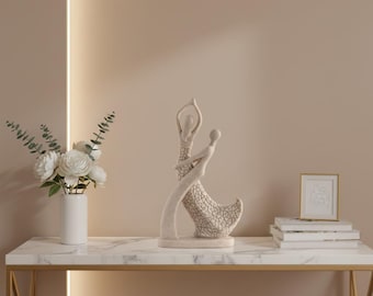 Op de afbeelding: Een crèmekleurige sculptuur van twee dansende figuren, geplaatst op een tafel met marmerlook en een gouden frame. Een witte vaas met witte bloemen en een ingelijste foto staan ook op de tafel. De achtergrond is neutraal beige.