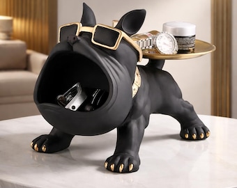 Op de afbeelding: Een zwart bulldogbeeldje met gouden accenten, met een goudkleurige zonnebril. De open mond van de bulldog dient als opbergruimte en een klein gouden dienblad op zijn rug houdt een horloge en een pot vast. De poten en halsband zijn goudkleurig.