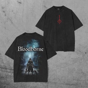 May include: Two black t-shirts. One features a red symbol, the other has the word "Bloodborne" and a graphic of a figure in a dark alley. The shirts have a vintage, worn look.