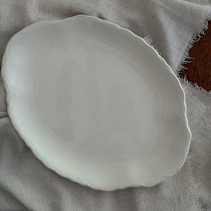 May include: An oval-shaped white ceramic serving platter with a smooth, glossy finish and a gently scalloped, wavy edge. The platter features a simple, clean design with subtle embossed detailing along the curved rim, providing a classic look.