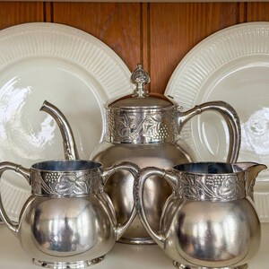 May include: A silver tea set with a teapot, two pitchers, and two white plates. The teapot and pitchers have a decorative grape and leaf design around the top. The plates have a ridged pattern.