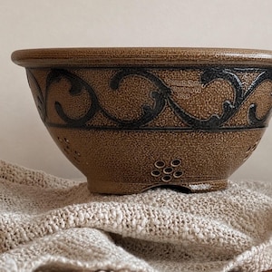 May include: A brown ceramic bowl with a textured surface, featuring a dark decorative scroll pattern around the upper rim and small flower-shaped perforations near the base. The text Wisconsin Pottery Columbus WI 1989 is stamped on the side.