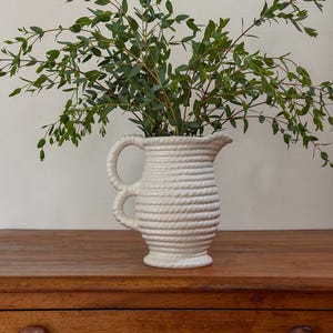 May include: A white ceramic pitcher with a rope-like texture, holding green leafy branches. The pitcher has two loop handles and sits on a wooden surface. The overall aesthetic is rustic and natural.