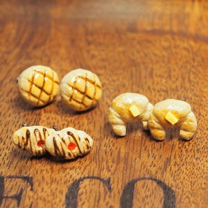 May include: Miniature food earrings in the image include two pairs of croissants with butter, two pairs of round buns with a crosshatch pattern, and two pairs of small pastries with chocolate drizzle and red dots. The earrings are on a wooden surface.