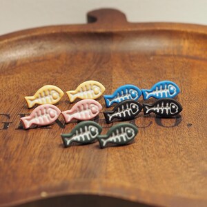 May include: A selection of colourful fish-shaped embellishments in yellow, pink, blue, black, and green. Each fish has a white bone design. The embellishments are arranged on a wooden tray.