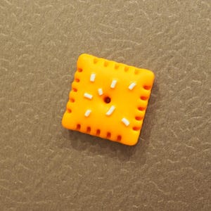 May include: A square orange cracker-shaped charm with rounded edges and a scalloped border. The surface features a small central hole and several short, raised white rectangular accents scattered across the top, mimicking a snack food item.