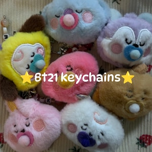 May include: A collection of seven plush BT21 keychains featuring various characters with embroidered facial expressions and pacifiers in colors including blue, yellow, pink, purple, brown, and white, with the text "BT21 keychains" in the center.