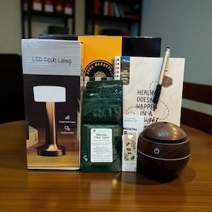 Può includere: Una collezione di articoli per la cura di sé su un tavolo di legno. Include una lampada da scrivania a LED, quaderni, bustine di Matcha Chai Latte, una penna, un piccolo diffusore di oli essenziali e una piccola scatola. La scatola della lampada dice "LED Desk Lamp".