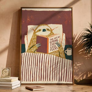 May include: A framed wall art print featuring a sloth lying in bed under a striped blanket, reading a book titled The Art of Giving Even Less Fucks. The sloth is positioned against a white pillow with a small coffee mug visible on the side.