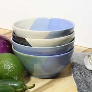 May include: A stack of four ceramic bowls featuring a color-blocked design in shades of light blue, cream, and dark charcoal. The bowls have a smooth, glossy finish and a rounded shape, presented alongside a red onion, two limes, and two peppers.