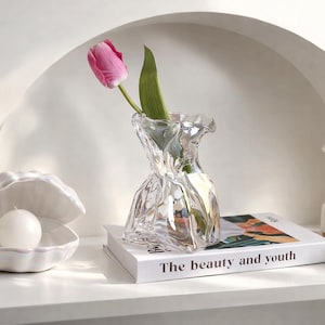 Może przedstawiać: Przezroczysty szklany wazon z różowym tulipanem i zieloną łodygą. Wazon stoi na książce zatytułowanej "The beauty and youth". Po lewej stronie biała muszla z białą świecą.