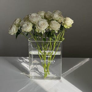 Op de afbeelding: Een heldere, rechthoekige glazen vaas gevuld met water en witte rozen. De vaas heeft de tekst "Un livre sur les Fleurs, A book about flower". De rozen staan in volle bloei, met groene stelen en bladeren.