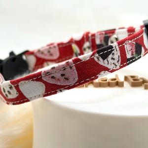 May include: A red fabric pet collar featuring a repeating pattern of white and black sushi rolls. The collar includes a black plastic buckle, a small silver-toned metal bell, and white stitching along the edges of the fabric strap.