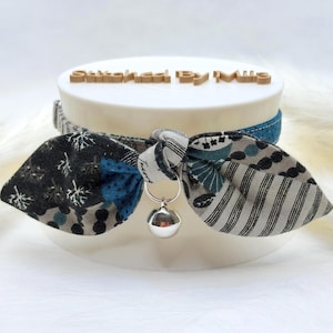 May include: A pet collar featuring a fabric bow tie with a multi-patterned design in shades of black, blue, beige, and white. The collar includes a small silver-toned metal bell attached to the centre of the bow. The text Stitched By Mia is visible.