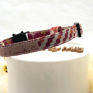 Puede incluir: Un collar para mascota con una correa de tela que presenta un patrón de círculos granates y naranjas sobre un fondo gris, acentuado con detalles de costuras blancas. El collar incluye una hebilla de plástico negro y un pequeño cascabel dorado.