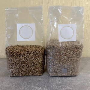 May include: Two transparent plastic bags filled with grains. The bag on the left contains light brown grains, while the bag on the right contains darker grains. Each bag has a white label with a circular design.