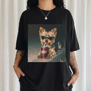 May include: Black t-shirt featuring a graphic of a Yorkshire Terrier wearing sunglasses and drinking from a Dr. Pepper can. The dog is brown, black, and tan. The Dr. Pepper can is red and white.