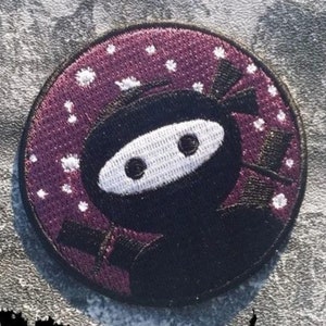 May include: A round purple and black embroidered patch featuring a cartoon ninja with a white mask and a black headband. The background is a dark purple with white stars.