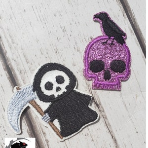 Op de afbeelding: Twee geborduurde strijkpatches met Halloween-thema's. Eén patch is een paarse glitter schedel met een zwarte kraai erop. De andere patch is een zwart-witte cartoon Grim Reaper die een zeis vasthoudt.