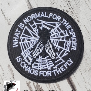 Pode incluir: Etiqueta bordada preta e branca com uma teia de aranha e uma mosca no centro. O texto ao redor da borda diz "O que é normal para a aranha é o caos para a mosca."