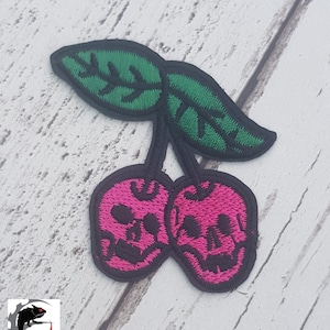 Op de afbeelding: Roze schedel kersen met groene bladeren. De kersen zijn geborduurd met zwarte contouren en de bladeren zijn geborduurd met zwarte contouren.