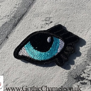 May include: Black and white embroidered eye patch with a turquoise blue iris. The eye patch is a whimsical and decorative accessory.
