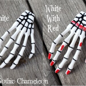May include: Two white plastic skeleton hand hair clips. One clip is all white with black details. The other clip has red nail polish on the fingertips.