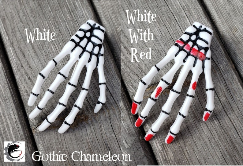 May include: Two white plastic skeleton hand hair clips. One clip is all white with black details. The other clip has red nail polish on the fingertips.