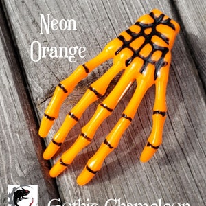 May include: Neon orange skeleton hand hair clip. The hand is made of plastic and has black details.