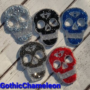 Könnte beinhalten: Fünf Strass-Schädel-Patches in Silber, Schwarz, Blau, Grau und Rot. Jeder Schädel hat ein detailliertes Design mit funkelnden Verzierungen. Der Text "GothicChameleon" befindet sich unten.