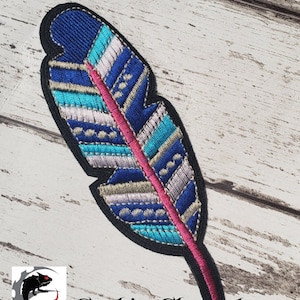 Può includere: Una piuma con motivo blu, turchese e bianco con una linea centrale rosa. La piuma è ricamata e ha un contorno nero.