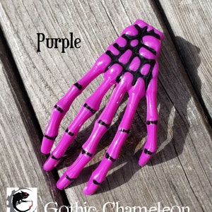 May include: Purple skeleton hand hair clip with black detailing. The clip is made of plastic and has a shiny finish.