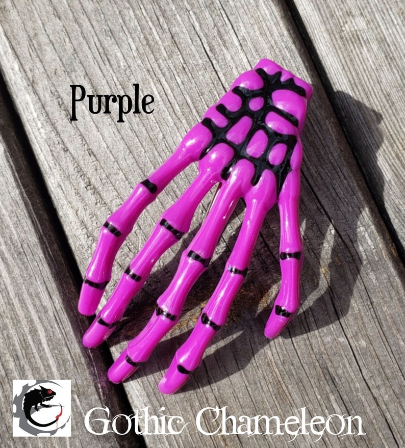 May include: Purple skeleton hand hair clip with black detailing. The clip is made of plastic and has a shiny finish.