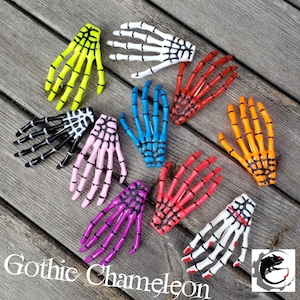 May include: A set of ten skeleton hand hair clips in various colors, including yellow, white, blue, red, orange, pink, and purple. The clips are made of plastic and have a black spiderweb design.