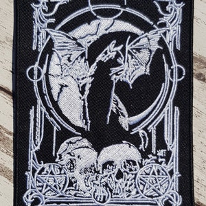May include: Black embroidered patch featuring a white design of a bat, skulls, and pentagrams. The design is framed by a circle and a square.