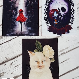 以下が含まれることがあります： 暗い森の中を歩く赤いフードをかぶった人物、ゴシック調の額縁に入ったカップル、そして頭に花をつけた白い猫の3つのデジタルアートプリントです。