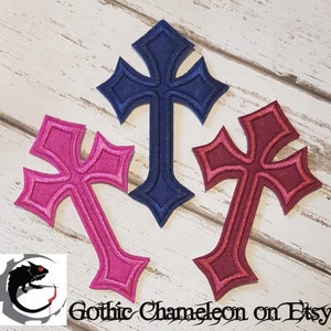 May include: Three embroidered iron-on patches in the shape of gothic crosses. The crosses are navy blue, pink, and burgundy.