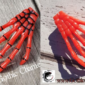 May include: Two plastic skeleton hand hair clips. One is red with black details, the other is solid red. Both are on a wooden surface.