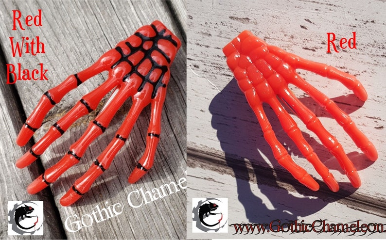 May include: Two plastic skeleton hand hair clips. One is red with black details, the other is solid red. Both are on a wooden surface.