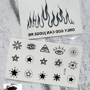 Puede incluir: Dos hojas de tatuajes temporales. La hoja superior presenta un diseño de llama negro y azul con el texto "ONLY GOD CAN JUDGE ME". La hoja inferior presenta una variedad de diseños en blanco y negro, incluyendo estrellas, ojos y un triángulo con un ojo dentro. El texto "K-136" es visible en la hoja inferior.