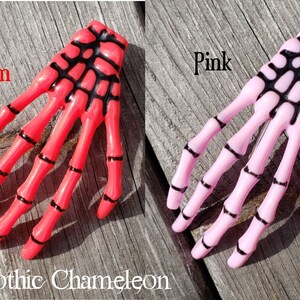 May include: Two skeleton hand hair clips, one in red salmon and one in pink. The clips have black detailing.