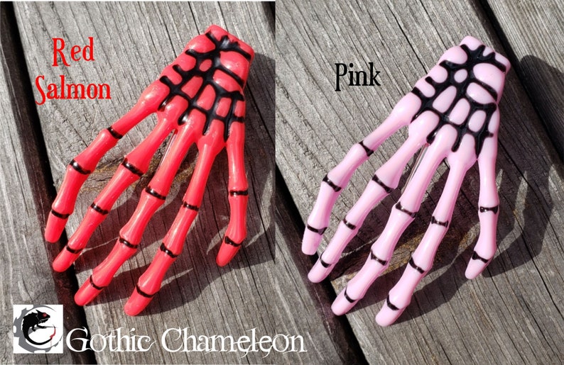 May include: Two skeleton hand hair clips, one in red salmon and one in pink. The clips have black detailing.
