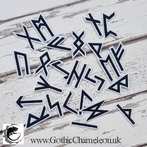 Puede incluir: Un conjunto de parches de runas bordadas en blanco y negro. Los parches presentan varios diseños angulares y geométricos, con un fondo blanco y costuras negras. Los parches están dispuestos sobre una superficie de madera de color claro. La dirección del sitio web www.GothicChameleon.uk es visible.