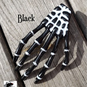 May include: Black skeleton hand hair clip with white details. The clip is on a wooden surface.