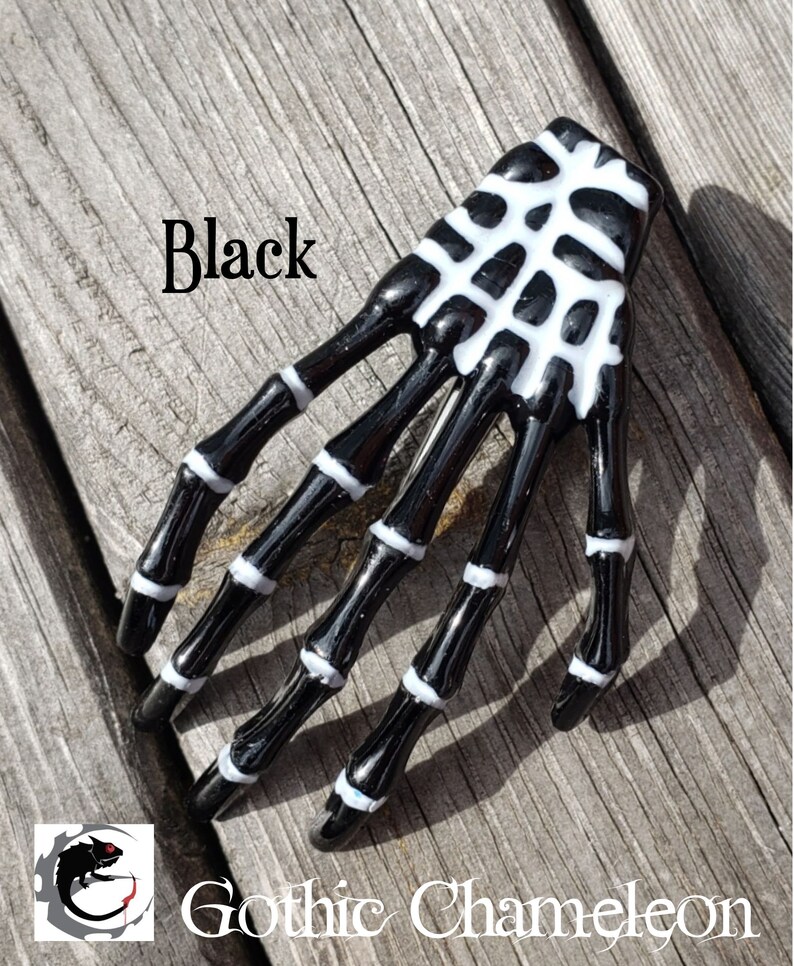 May include: Black skeleton hand hair clip with white details. The clip is on a wooden surface.