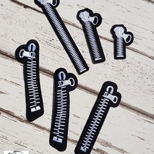 May include: Six black and white zipper patches of varying sizes. The zippers are white with black teeth and the zipper pulls are white.