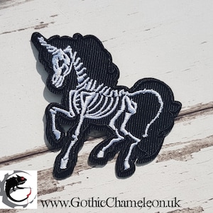 May include: Black and white embroidered patch of a skeleton unicorn. The unicorn is standing on all four legs with its head turned to the side. The unicorn's horn is visible. The patch is on a white wooden background.